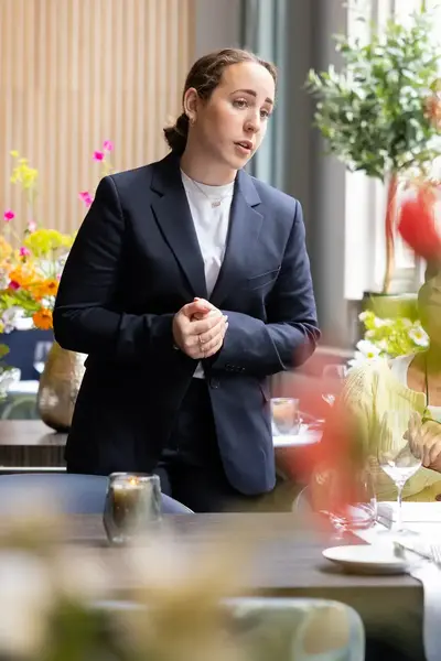 student in net pak staat aan tafel bij een gast in een restaurant met veel bloemen, tafeldecoratie en glazen.