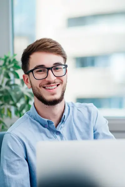Man met bril en baard, bruin haar en een glimlach, zit achter een bureau met een laptop