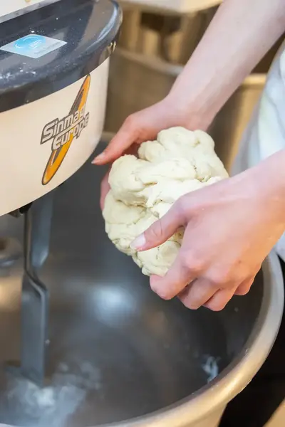 Close-up van handen in bakkerskleding die een deegbol vasthouden boven een deegmixer.