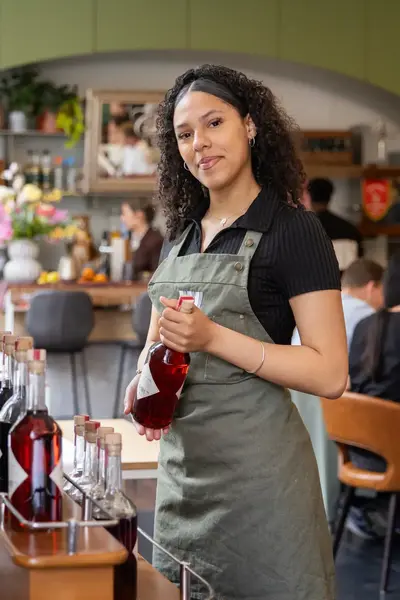 Een jonge vrouw in een groen schort houdt een fles drank vast in een restaurant. Op de achtergrond zitten mensen aan tafels.