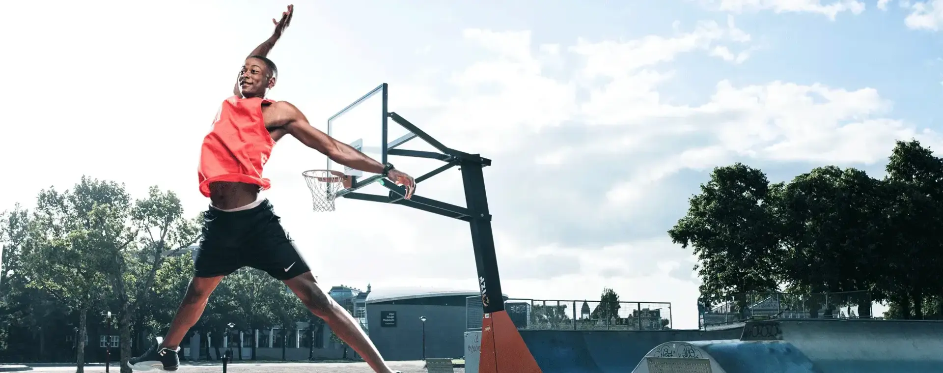 Basketballer maakt dunk beweging op den basketbalveld buiten. Je ziet een skatepark op de achtergrond.