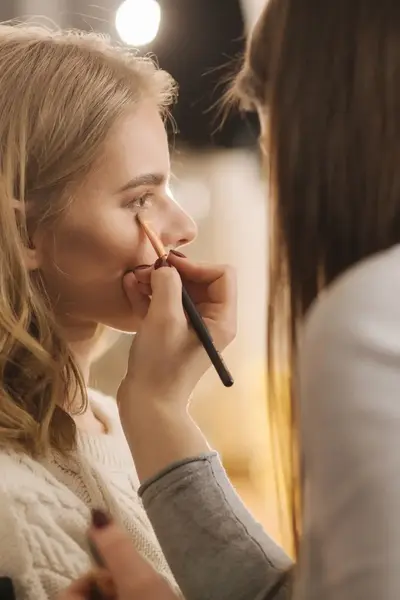 Visagist brengt make-up aan op het gezicht van een jonge vrouw in een professionele studio met zachte verlichting.