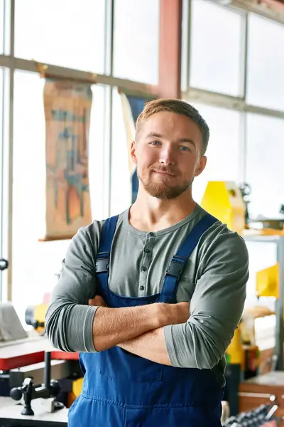 Jonge man met overall aan en over elkaar geslagen armen staat in een werkplaats of praktijklokaal en kijkt lachend in de camera.