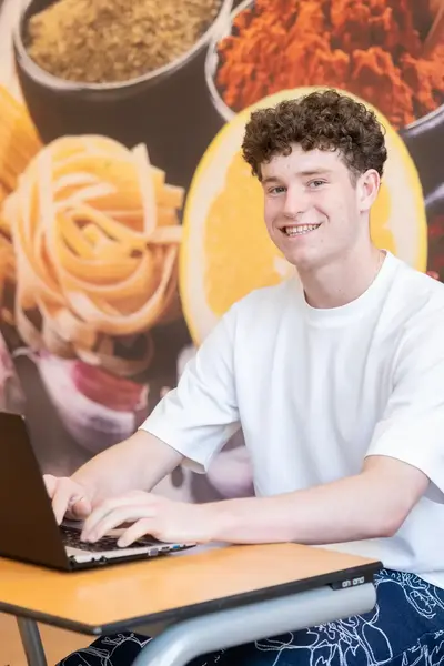 Een jonge man werkt op zijn laptop in een ruimte met een grote foto van eten op de muur.