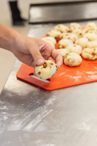 Close-up van een hand die een rozijnenbolletje vasthoudt boven een rode bakplaat vol deegbolletjes.