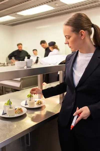 Een vrouw in pak controleert gerechten in een keuken met een stift in haar hand. Op de achtergrond zijn koks druk aan het werk.
