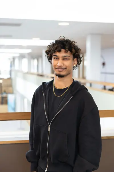 Student met krullen en zwarte hoodie staat in een lichte schoolgang en kijkt lachend naar de camera.