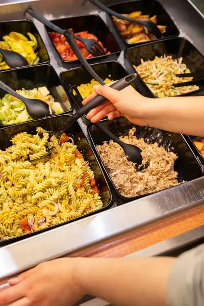 Medewerker schept pasta op bij een saladebar vol verse gerechten zoals groentesalades, pastasalade en fruit.