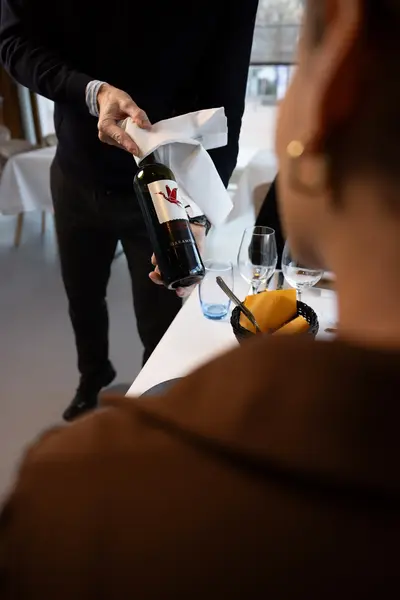 Een medewerker presenteert een fles wijn aan een gast in een restaurant. Op tafel staan glazen en bestek.