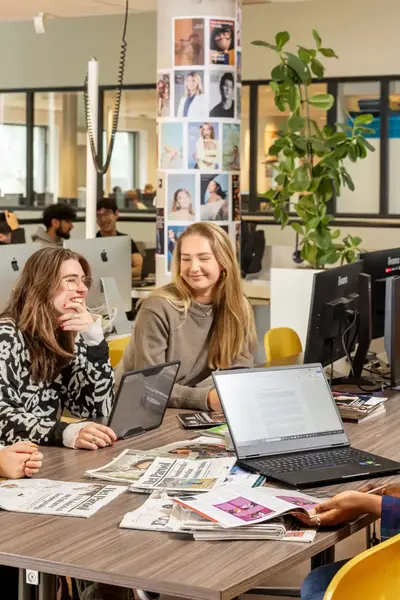 Vier studenten waarvan één achter een laptop zitten aan een tafel waarop verschillende dagbladen liggen.