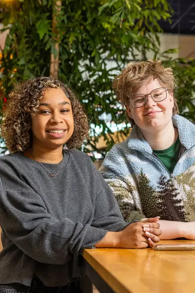 Twee studenten poseren lachend aan een houten tafel.