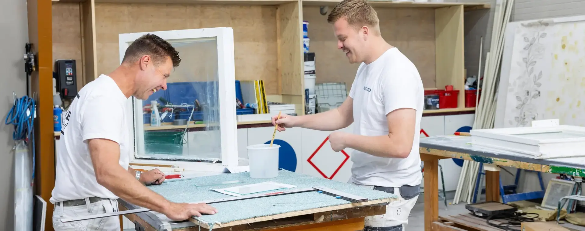Twee mannen in witte werkkleding lachen terwijl ze samen aan een schilderklus werken. Ze staan in een werkplaats en bereiden een kozijn voor.