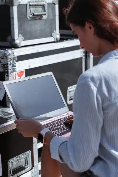 Vrouw werkt op laptop tussen flightcases op locatie, ter voorbereiding van een evenement of optreden.