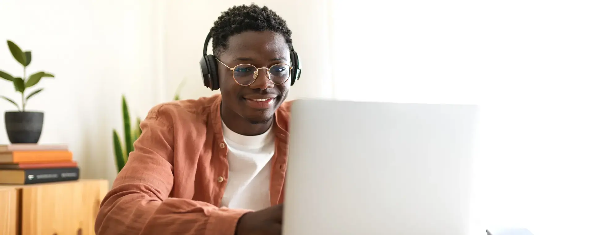 Student volgt online les achter zijn laptop. Hij draagt een koptelefoon en een bril, en zit glimlachend aan een bureau met boeken en een smartphone.