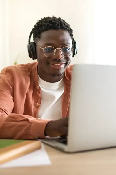 Student volgt online les achter zijn laptop. Hij draagt een koptelefoon en een bril, en zit glimlachend aan een bureau met boeken en een smartphone.