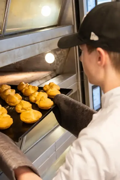 Student haalt een bakplaat met verse soesjes uit de oven tijdens de bakkersopleiding bij het ROC. Hij draagt een bakkersuniform en ovenhandschoenen.