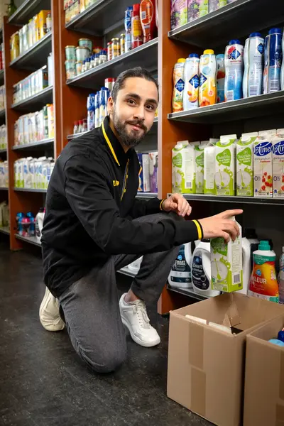 Winkelmedewerker vult schappen in een supermarkt, terwijl hij een pak yoghurt uit de doos pakt."