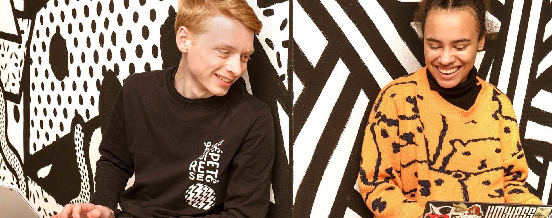 Twee studenten zitten met laptops lachend voor een grafische geschilderde muur
