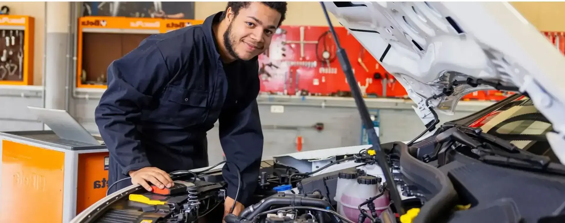 Student autotechniek werkt aan een auto in een werkplaats en kijkt lachend naar de camera.