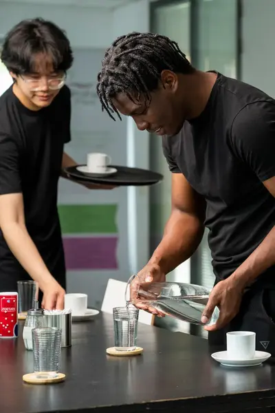 Twee jonge mannen dekken een tafel. De één schenkt water in een glas, terwijl de ander kopjes op een dienblad plaatst. Op de achtergrond staat een witte piano.