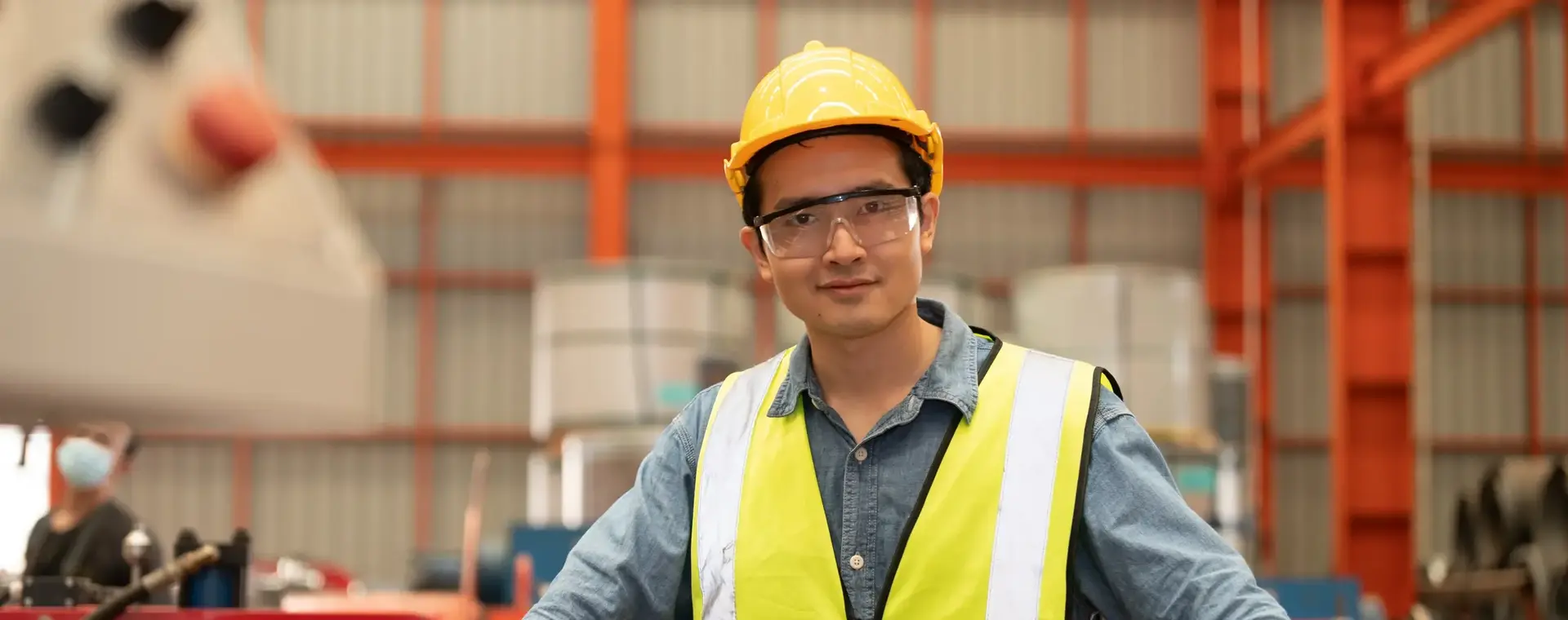 Man met gele helm en geel hesje tussen oranje machines
