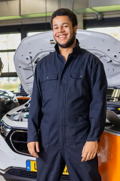 Student in overall staat lachend in een autowerkplaats voor een geopende motorkap.