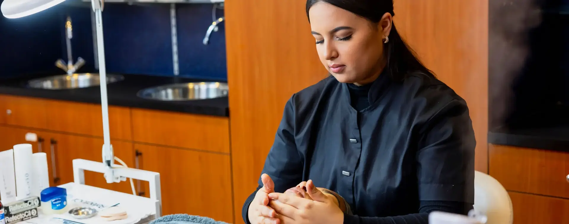 Student schoonheidsspecialist voert een gezichtsmassage uit bij een klant in een salonomgeving met handdoek, behandelstoel en verzorgingsproducten.