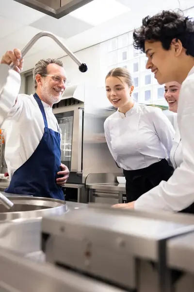 Een docent tilt lachend het deksel van een grote kookketel terwijl drie studenten in kokskleding aandachtig toekijken.