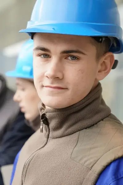 Vier mannen met bouwhelmen op staan op een bouwplaats. De voorste jonge man kijkt in de camera.