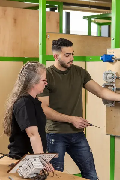 Student legt bedrading uit aan docent bij een oefeninstallatie met stopcontacten, schakelaars en elektriciteitskabels in een lokaal met groene kaders en houten panelen.