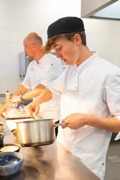 Een jonge kok in witte koksbuis en zwarte muts is geconcentreerd bezig met het bereiden van een gerecht in een pan, terwijl op de achtergrond een collega werkt.