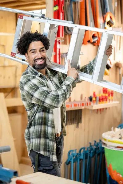Jonge man met geruite blouse in timmerwerkplaats houdt een ladder op zijn schouder en lacht in de camera.