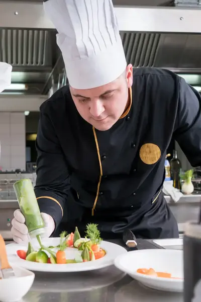 Een student kijkt aandachtig mee terwijl een chef-kok een gerecht zorgvuldig opmaakt met een groene saus.
