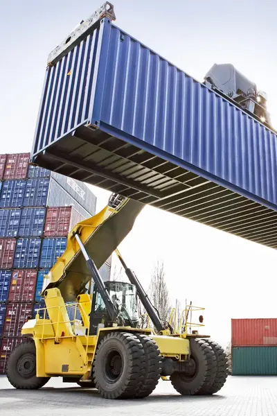 Gele heftruck tilt grote blauwe zeecontainer op tussen stapels containers op een overslagterrein.