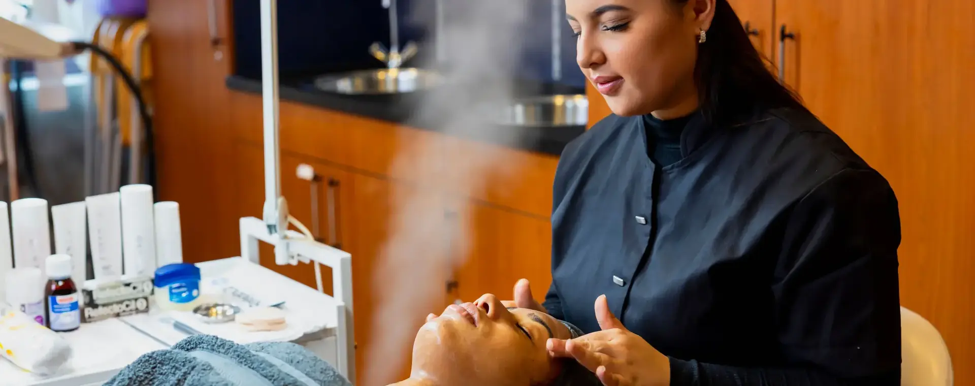 Student schoonheidsspecialist voert een gezichtsbehandeling uit bij een klant onder een gezichtsstomer, omringd door verzorgingsproducten in een salonsituatie.