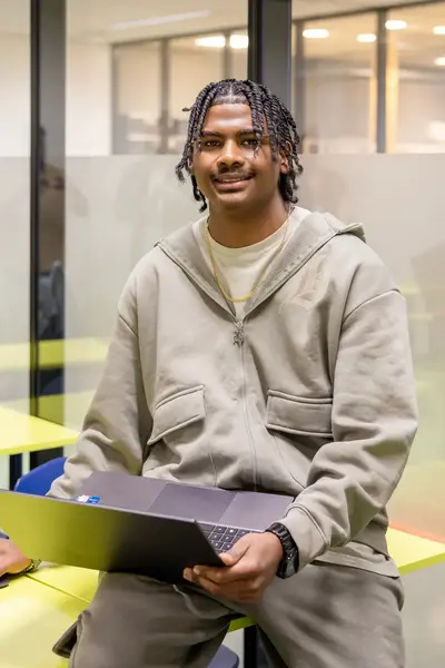 Een student zit in een klaslokaal met gele tafels en blauwe stoelen en werkt op een laptop.