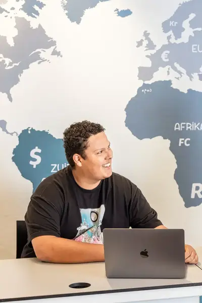 Een docent zit op een bureau in een ruimte met een wereldkaart aan de muur en voert een gesprek met een student.