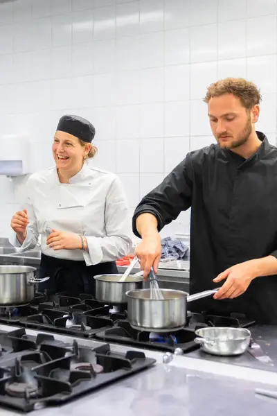 Drie koks staan samen in een . Eén is aan het roeren in een pan, terwijl de anderen in gesprek zijn. 