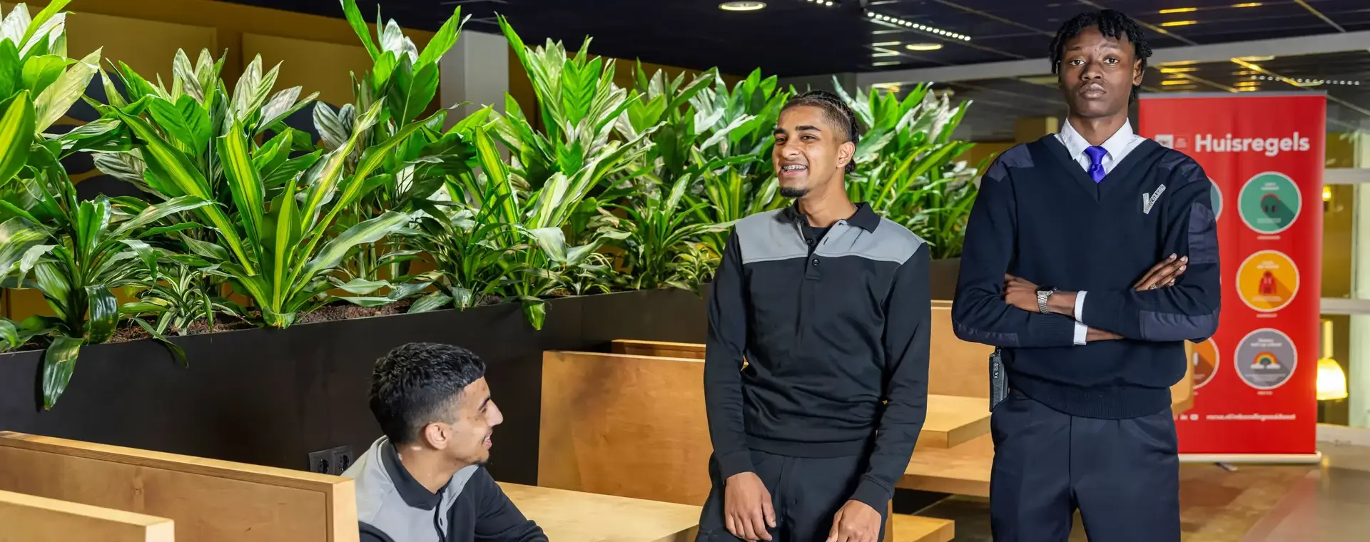 Drie studenten in beveiligingsuniform poseren in een schoolomgeving met groene planten en een bord met huisregels op de achtergrond.