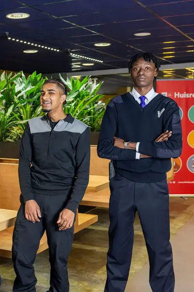 Drie studenten in beveiligingsuniform poseren in een schoolomgeving met groene planten en een bord met huisregels op de achtergrond.