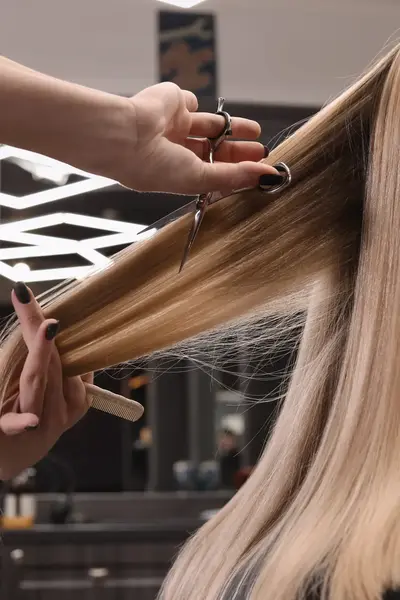 Een kapper knipt lang blond haar met een schaar en houdt het haar recht met een kam. De klant zit in een stoel in de salon.