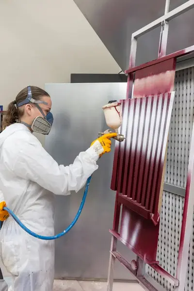 Student met een masker en overall aan spuit een coating op een paneel terwijl een docent met een masker op toekijkt en aanwijzingen geeft.