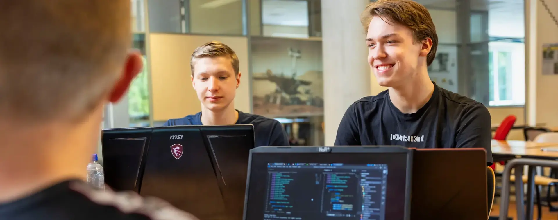 Vier studenten werken samen op laptops in een klaslokaal, met programmeercode zichtbaar op het scherm.