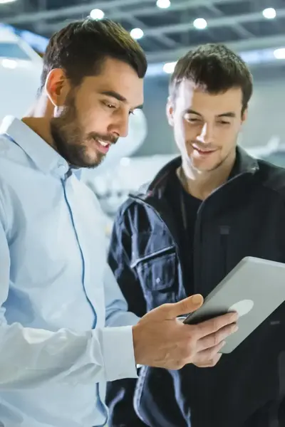 Man met blouse aan heeft Ipad in zijn hand en laat iets op zijn Ipad zien aan een andere man. Ze staan in een hangar met twee vliegtuigen op de achtergrond.