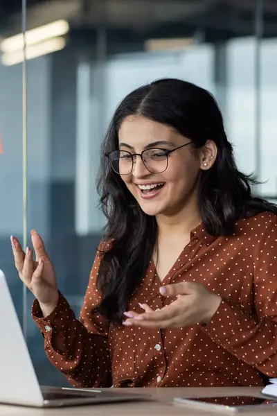 Vrouwelijke student met bril zit achter een laptop in een glazen kantoorruimte en reageert enthousiast tijdens een videogesprek. Achter haar hangen kleurrijke post-its op het raam.