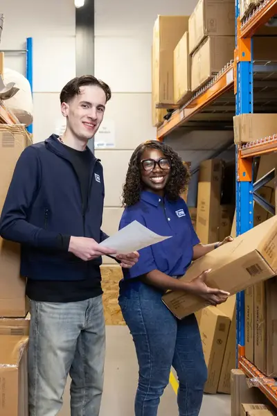 Twee studenten staan tussen stellingen vol dozen, één houdt een pakbon vast en de ander tilt een doos.