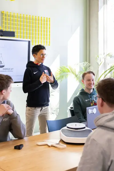 Vijf studenten zitten rondom een tafel en luisteren naar een docent die les geeft en daarbij het digibord gebruikt.