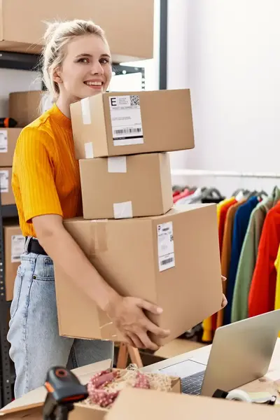 Vrouw in magazijn tilt meerdere dozen bij werkplek met laptop, kledingrek en verzendmaterialen op de achtergrond.