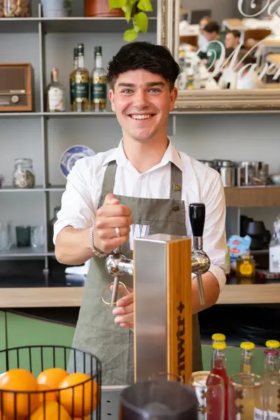 Jonge man in wit overhemd en schort tapt een drankje achter de bar. Op de achtergrond staan flessen, servies en een spiegel met tekst.