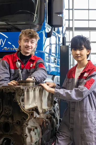 Twee studenten in overall werken aan een motorblok in een garage.